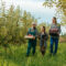 Two bearded mustachioed farmer field workers go and tell a woman about harvesting her tablet. Apple orchard, agricultural business, smiles on faces, success at work. Workers in the apple garden.